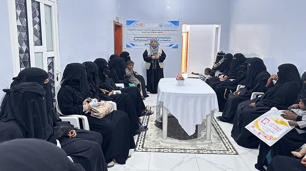 A group of women are seated around a table inside a room, listening to a woman in a UNFPA vest standing and speaking at the head of it