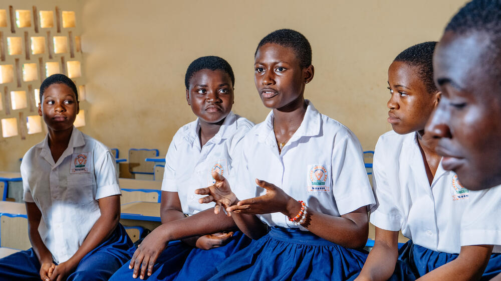 Quatre jeunes filles sont assises sur un banc, dans une salle de classe bien éclairée. Elles portent des uniformes scolaires comportant une chemise blanche et une jupe bleue. Une jeune fille, au centre, parle avec animation