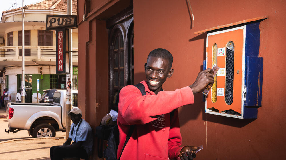 Un joven con sudadera roja sonríe ampliamente mientras sostiene condones frente a un dispensador de condones montado en la pared.