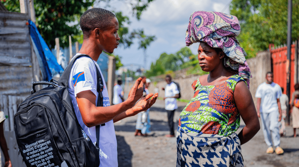  A young man in a white t-shirt with a black backpack speaks to a pregnant woman carrying a package on her head, as they stand in the street