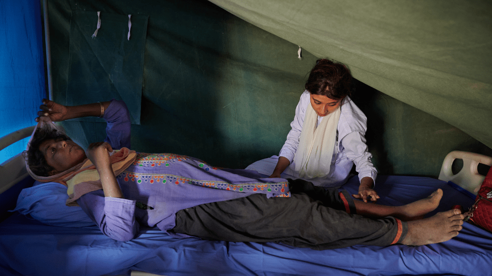  A pregnant woman in a purple tunic lies on a hospital bed in a green tent. A woman sits on the bed beside  her, checking her legs for signs of edema.