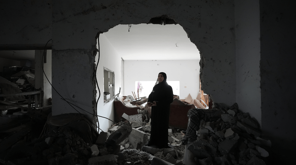 A woman dressed in black stands in the rubble of her home.