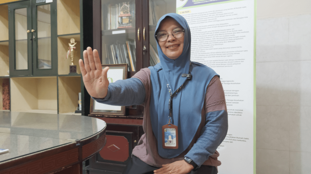 A woman in a blue head covering and glasses smiles as she holds her hand palm-up towards the camera