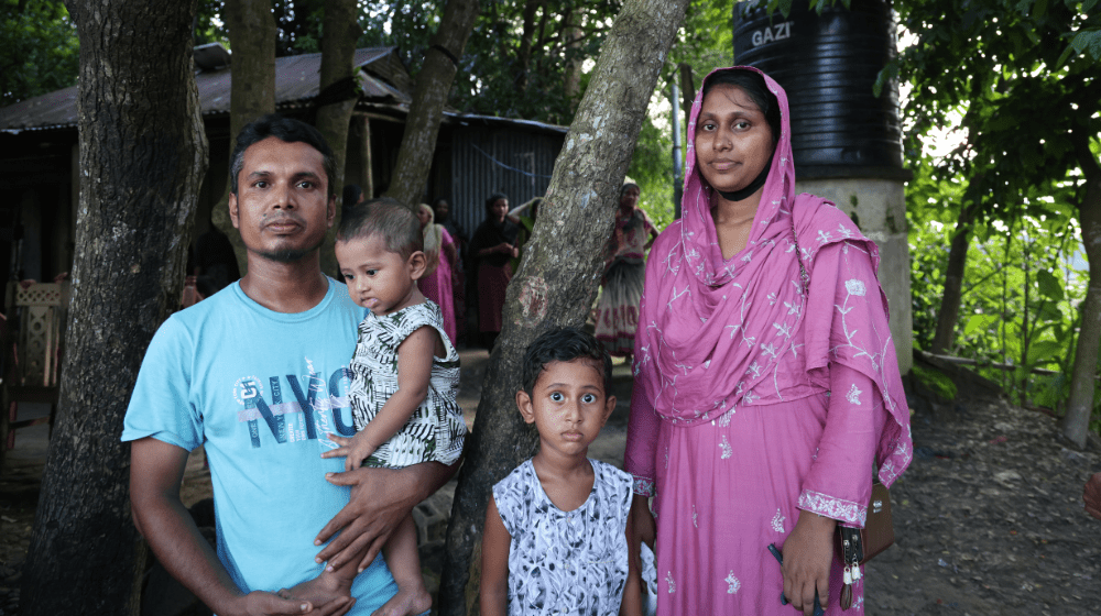  A man and woman stand with two young boys in front of a tree and a wooden house.
