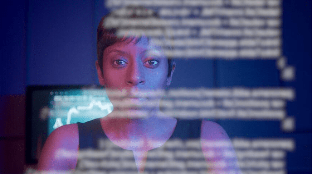 A woman is seen reading white text printed on transparent glass. She is viewed through the glass and is partly concealed by the text. Behind her is a computer monitor showing a chart
