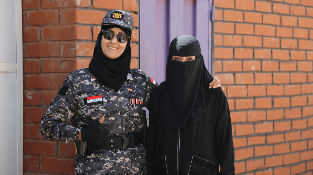 A woman in a security uniform wearing a cap, black hijab and dark sunglasses smiles broadly and gives the thumbs up, with her left arm around the shoulders of another woman dressed in a black niqab
