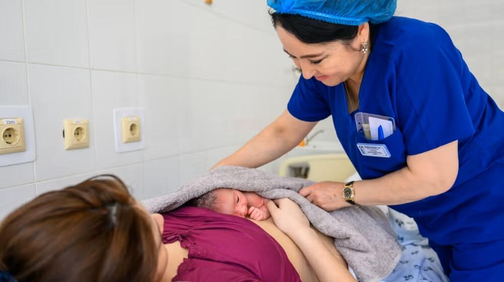 عاملة صحية ترتدي زيًا أزرق وشبكة شعر تضع مولودًا جديدًا ملفوفًا ببطانية رمادية على صدر أم ترتدي قميصًا أرجوانيًا في غرفة مستشفى مشرقة ذات أرضيات من البلاط.