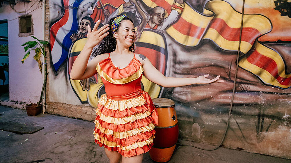 Une jeune femme en tenue de scène orange et jaune danse devant une fresque représentant un groupe de percussionnistes.
