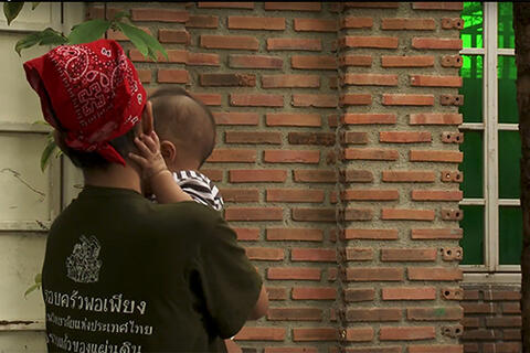 Mother wearing black t-shirt and red bandanna holds baby while standing outside brick structure