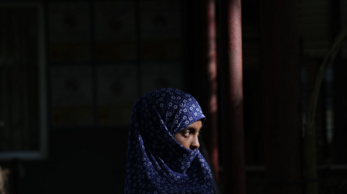 Woman wearing a blue hijab sits in a dimly lit room.
