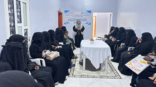 A group of women are seated around a table inside a room, listening to a woman in a UNFPA vest standing and speaking at the head of it