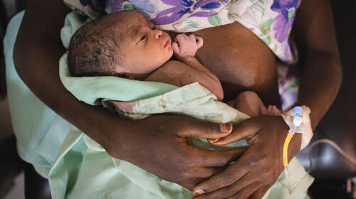 An adolescent girl holds her newborn daughter in a bloodied blanket. There is an IV line in the girl’s wrist.