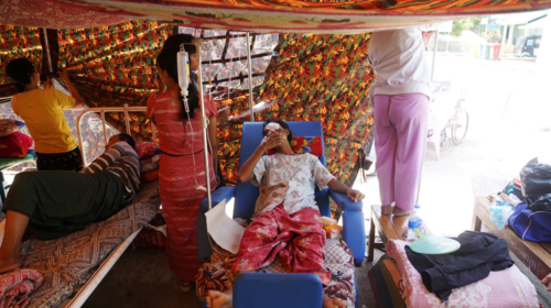 A woman lies on a hospital bed inside a makeshift tent, beside a man also on a bed and three women hanging up material to shade them from view