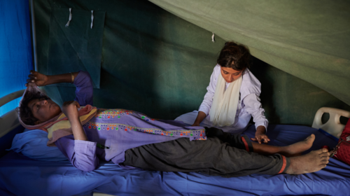  A pregnant woman in a purple tunic lies on a hospital bed in a green tent. A woman sits on the bed beside  her, checking her legs for signs of edema.