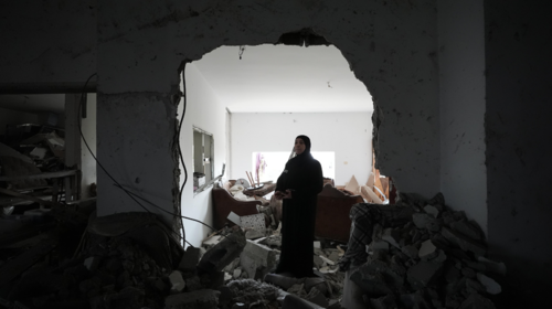 A woman dressed in black stands in the rubble of her home.