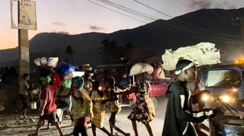 On a road with cars at trucks, people flee in near darkness carrying belongings on their heads, mountains visible in the background