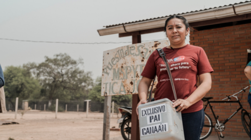 A woman in a red t-shirt stands in front of a building holding a container.