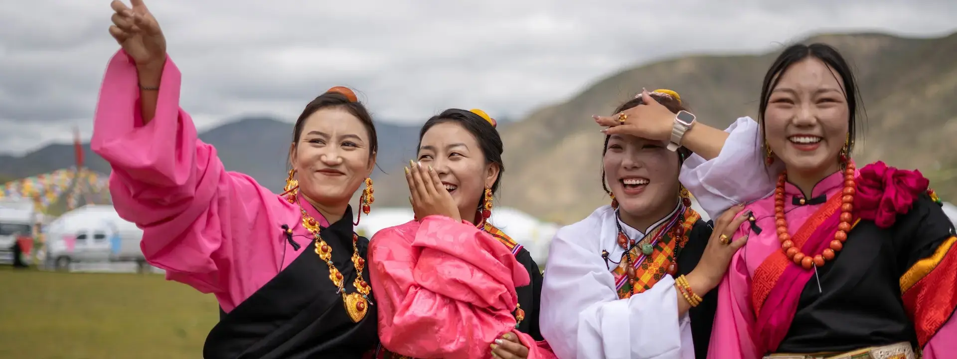 Four young women are adorned in traditional clothing and jewelry and stand together smiling in front of a mountainous landscape.