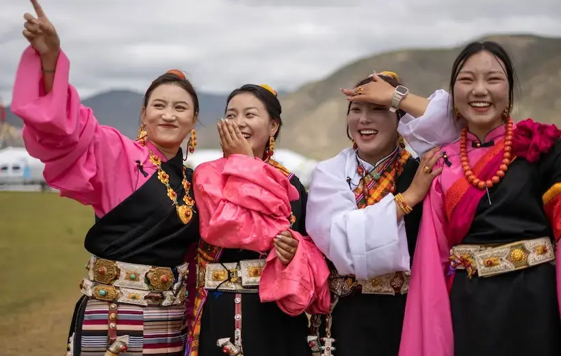 Four young women are adorned in traditional clothing and jewelry and stand together smiling in front of a mountainous landscape.