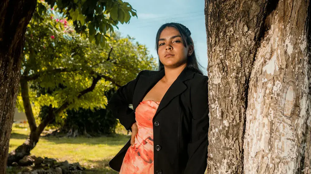 A young girl wearing a dress and blazer is leaning on a tree and confidently looks towards the camera.