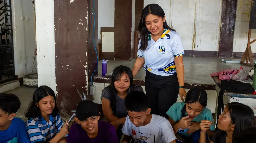 A young woman in a white polo shirt smiles and stands behind a group of young people who are sitting down