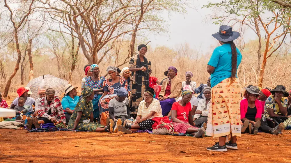 A woman in a blue t-shirt and a sunhat with the UNFPA logo stands before a crowd of women sitting and standing under trees