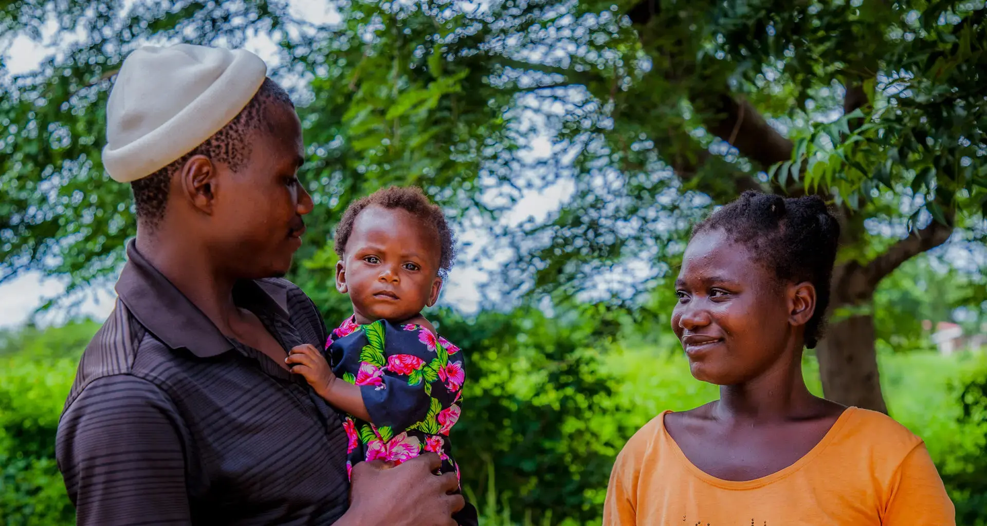 A father holds his infant in his arms and looks towards his smiling wife. 